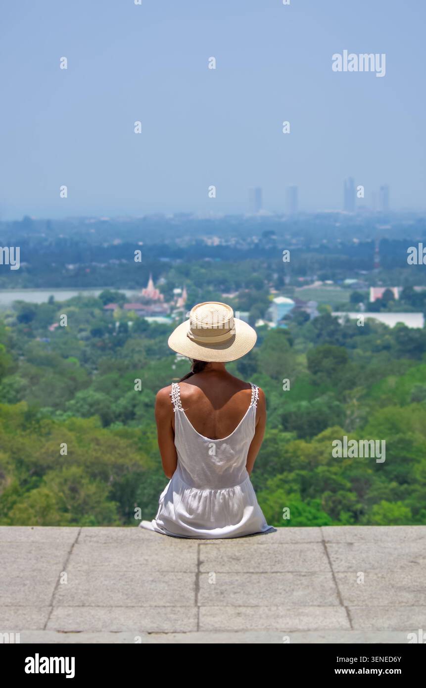 Eine junge Frau in weißem Kleid und Strohhut sitzt auf einem Steinberg mit Blick auf eine riesige tropische Stadtlandschaft mit üppigem Grün, Tempeln und einem Stockfoto