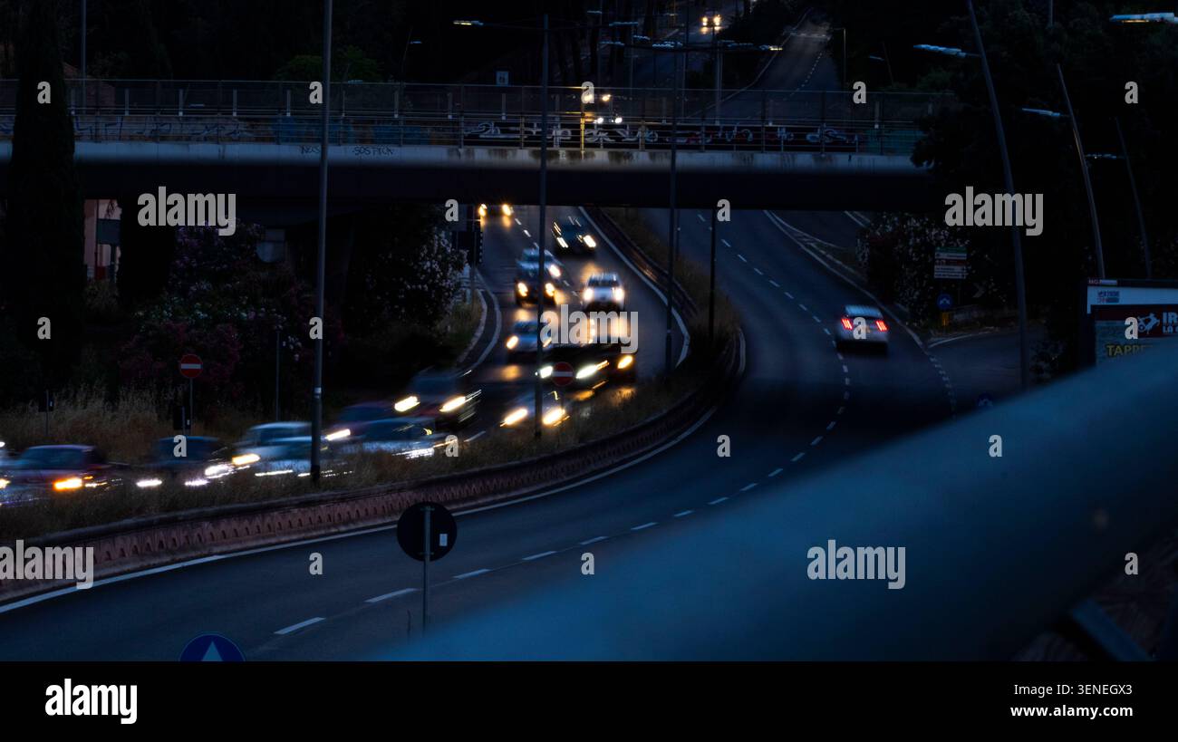 Der abendliche Verkehr fließt unter einer Überführung in Rom, Italien. Die Scheinwerfer beleuchten eine kurvige Stadtstraße zur blauen Stunde und schaffen eine stimmungsvolle Stadtlandschaft mit Stockfoto
