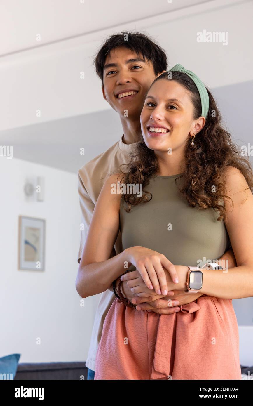 Paare umarmen und lächeln, während sie im Wohnzimmer mit olivfarbener Oberseite, Smartwatch und gerahmtem Druck stehen Stockfoto