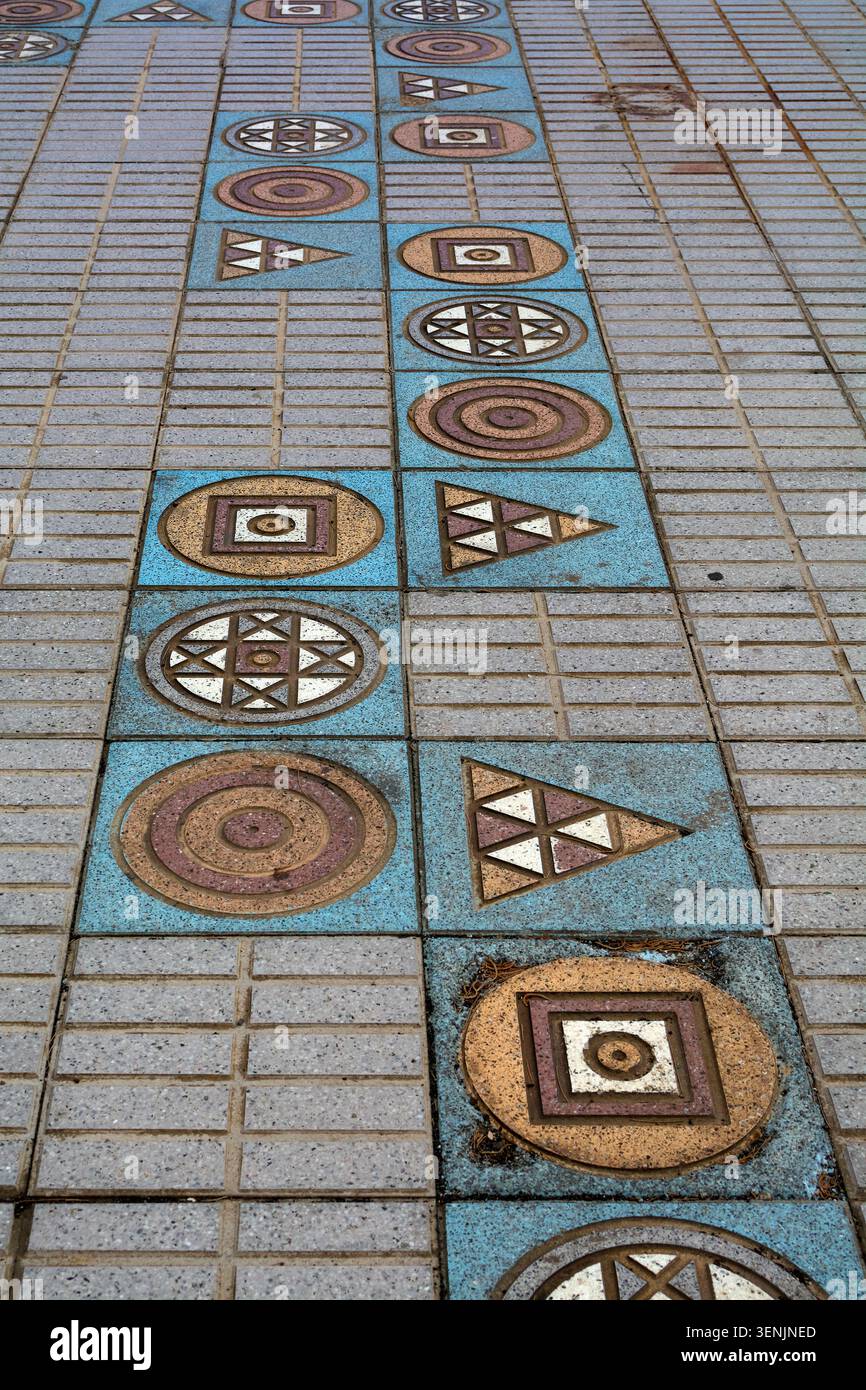 Ein geometrischer Mosaikweg aus Kreisen, Quadraten und Dreiecken in Blau- und Erdtönen ist in einen Fußgängerweg mit Steinfliesen eingebettet. Stockfoto