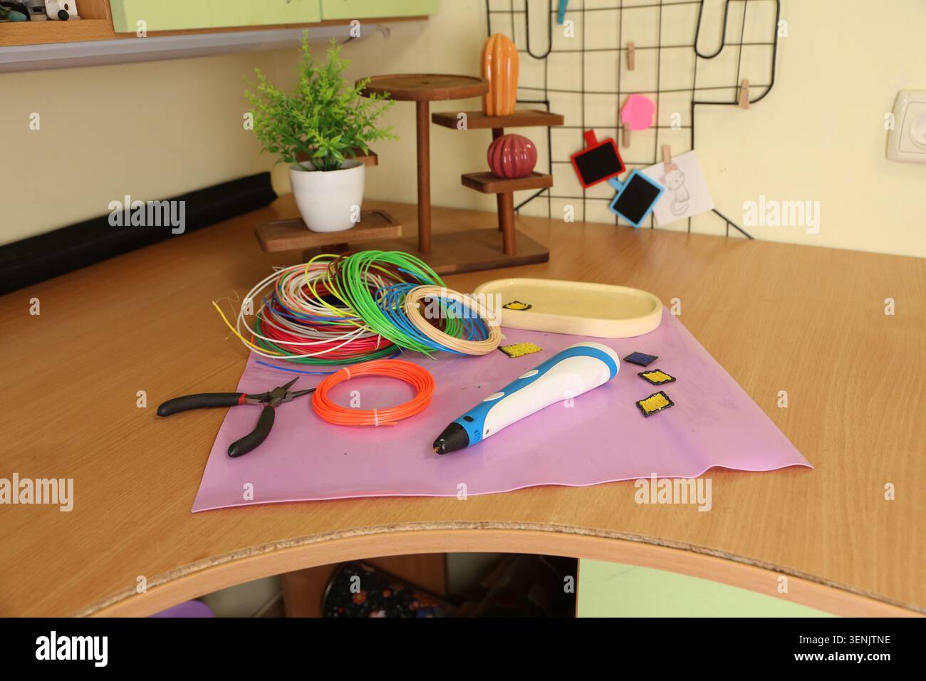 Ein Arbeitsbereich wird mit verschiedenen Gestaltungswerkzeugen eingerichtet. Es gibt einen 3D-Druckstift, bunte Drähte, Scheren und Fächer. Der Bereich hat eine grüne Pflanze und dekorative Gegenstände im Hintergrund. Stockfoto