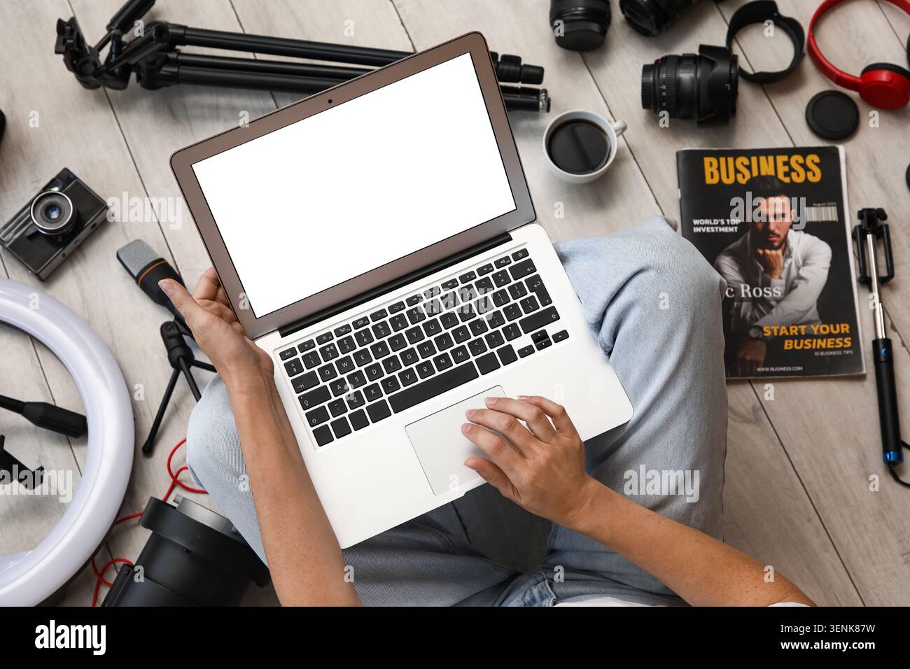 Person mit Laptop mit leerem Bildschirm, umgeben von Vlogging- und Fotoausrüstung auf Holzboden Stockfoto