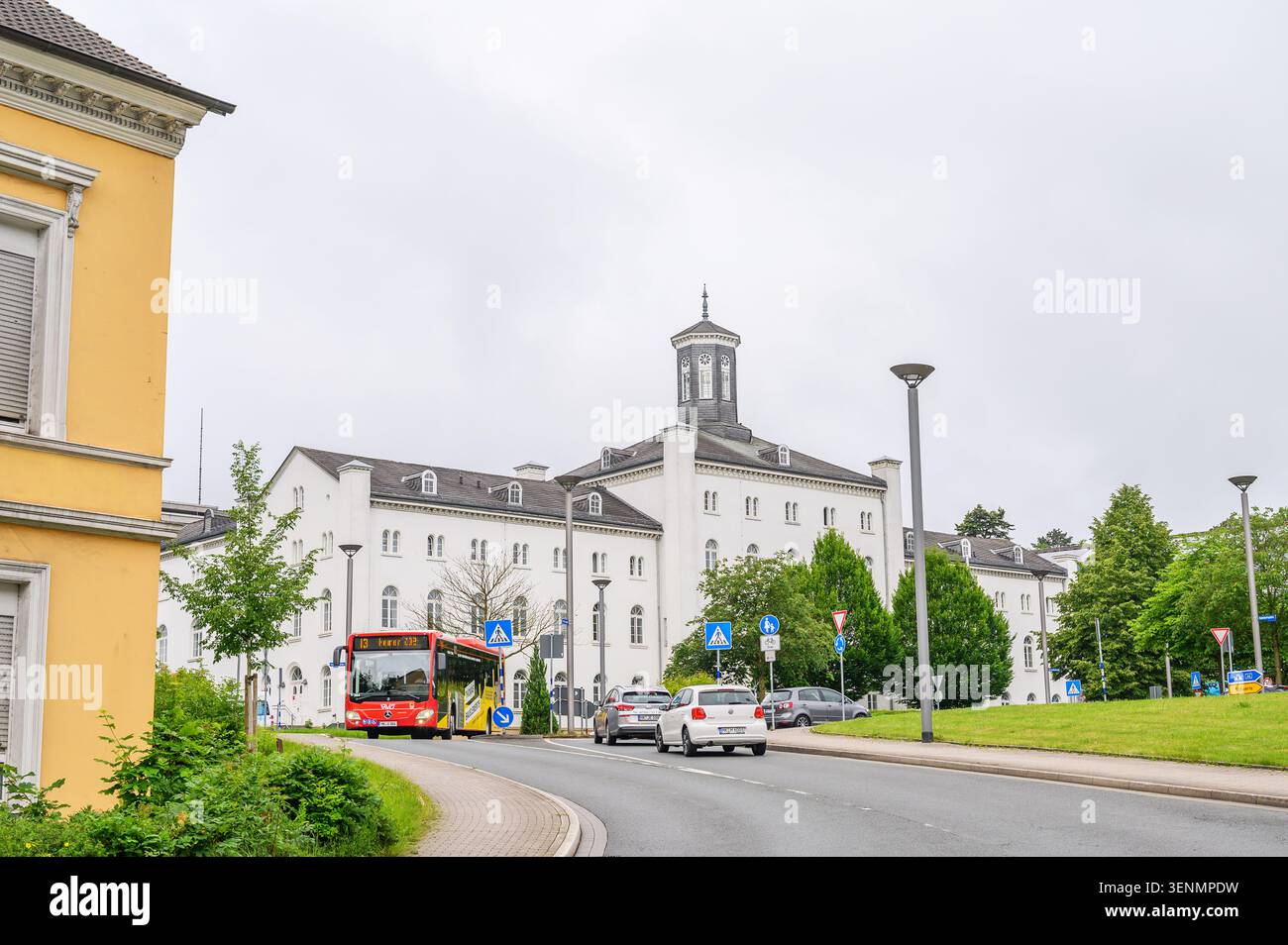 Iserlohn, Deutschland - 05. Juni 2026: Straßenblick mit rotem öffentlichem Bus vorbei an historischem Gebäude unter bewölktem Himmel. Stockfoto