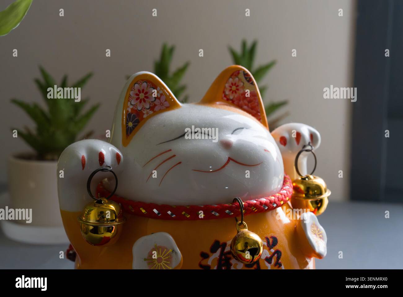 Keramikkatzenfigur mit goldenen Glocken und rotem Kragen. Die Katze hat geschlossene Augen und einen lächelnden Ausdruck. Die Pfoten halten kleine, glänzende Glocken. Grün p Stockfoto