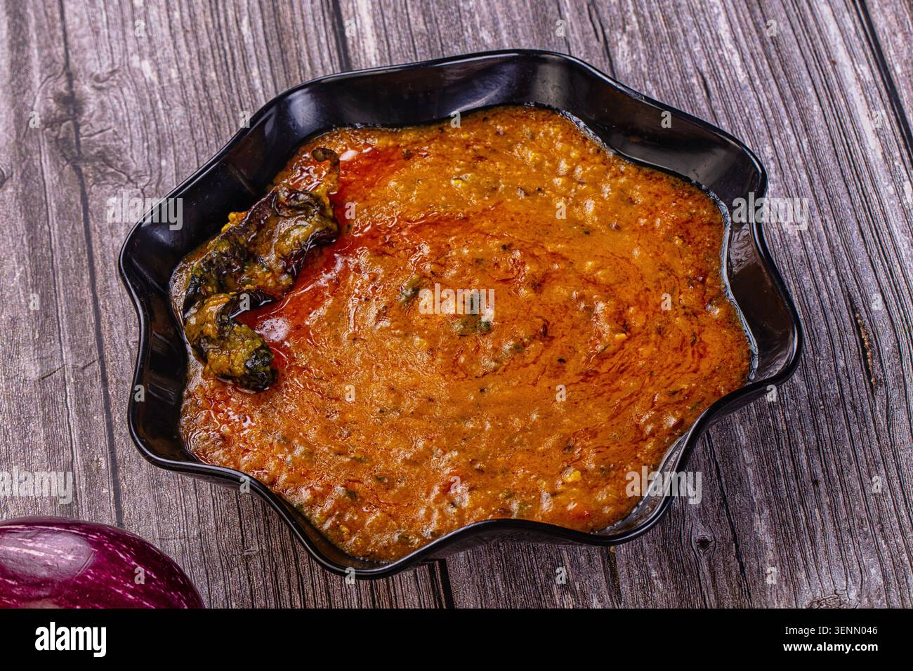 Nahaufnahme des traditionellen indischen Dal Tadka Linsencurry. Scharfes rot-gelbes Gericht in schwarzer Schüssel auf rustikalem Holztisch mit frischer Aubergine und o Stockfoto