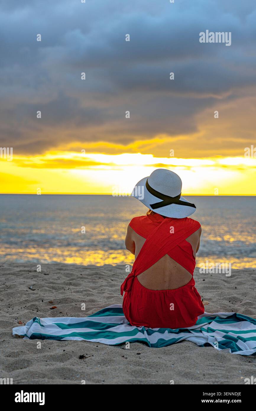 Frau mit Hut, die den Sonnenuntergang am Strand beobachtet Stockfoto