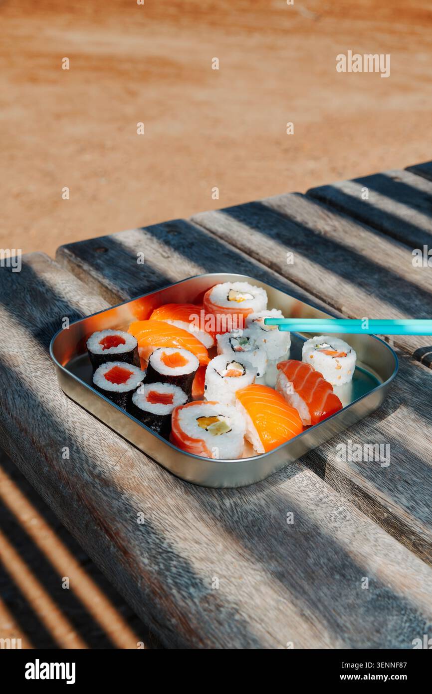 Mit leerem Platz über der Szene füllt eine Auswahl an Sushi und Nigiri eine Lunchbox aus Metall auf einer Holzbank, während Essstäbchen auf ein R zeigen Stockfoto