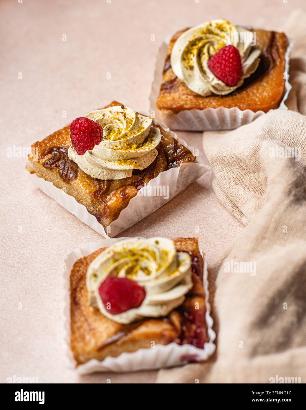 Drei quadratische Frangipan-Torten mit Schlagsahne, Pistazienstaub und frischen Himbeeren, die auf einer rustikalen Oberfläche angeordnet sind. Stockfoto