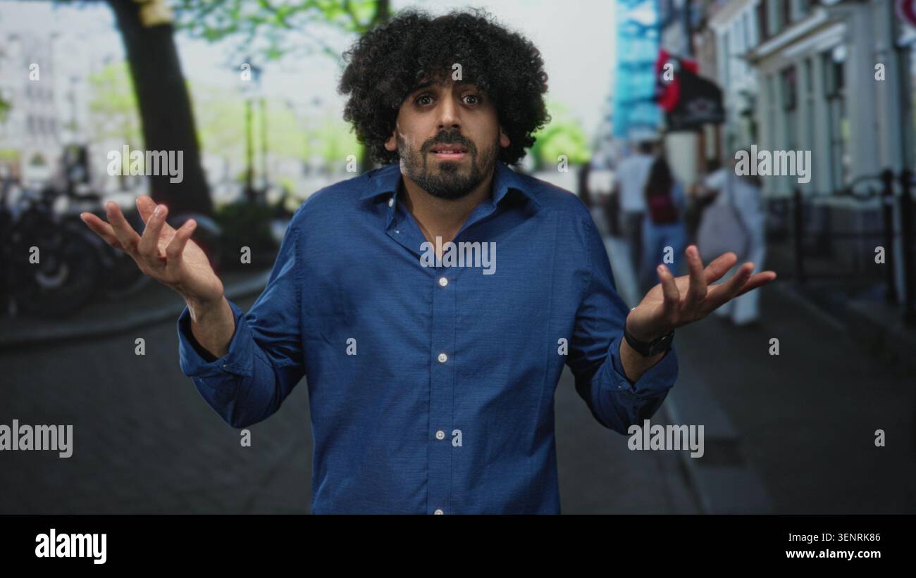 Mann mit lockigen Haaren, Sträucher mit offenen Händen auf einer Stadtstraße, blaues Hemd, sichtbare Palme, umgeben von verschwommenen Fußgängern und Fahrrädern; Verwirrung. Stockfoto