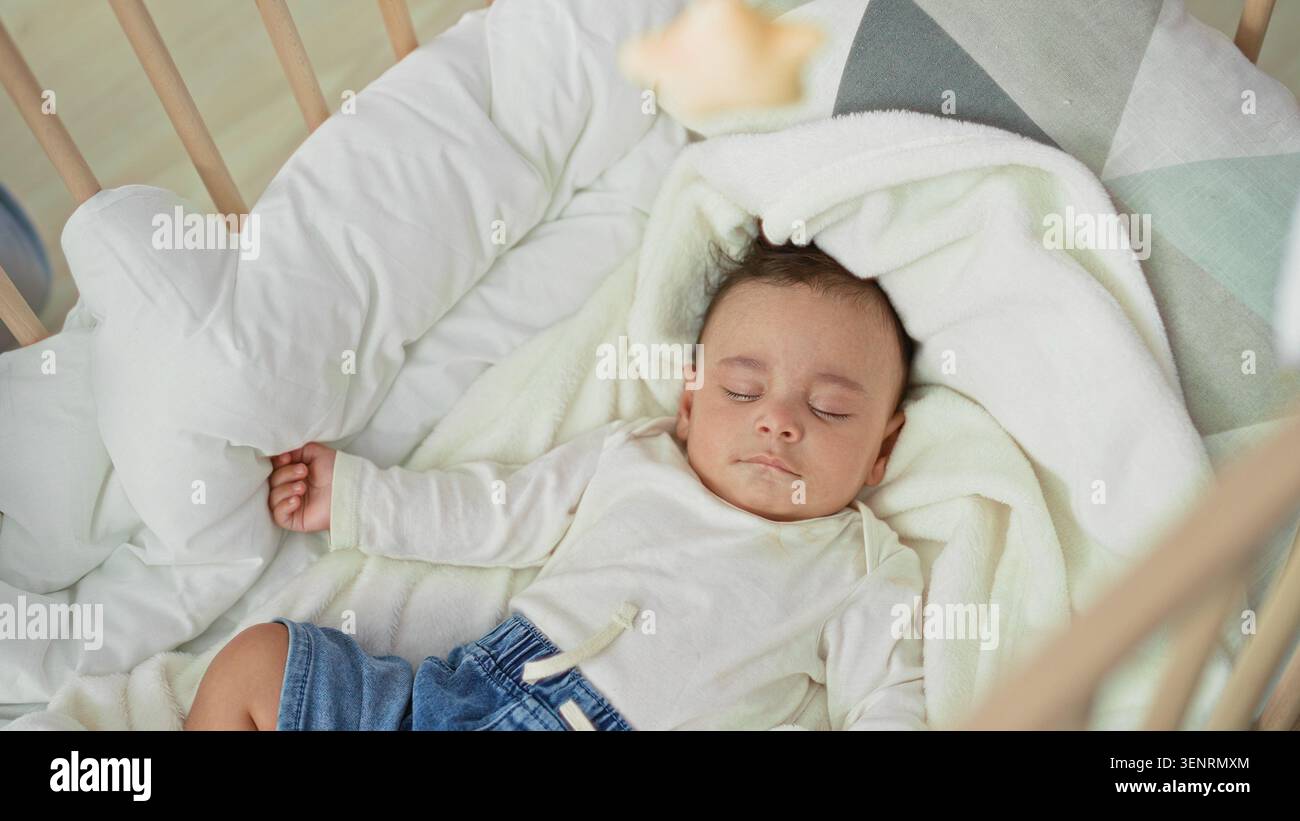 Hispanischer Kleinkind, der auf dem Rücken in einem hölzernen Wiegenbett mit weißer Decke und Jeansshorts schläft, Hand auf Kissen im Kinderzimmer gerollt; friedliche Ruhe. Stockfoto