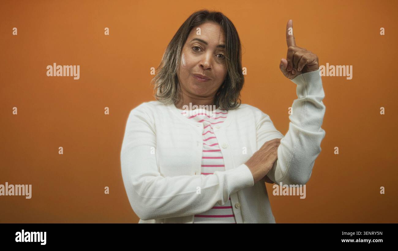 Die Frau zeigt den Zeigefinger nach oben, trägt im Studio eine weiße Strickjacke und ein gestreiftes Oberteil. Stockfoto