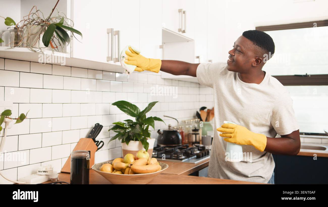 Lächelnder junger Mann, der Küchenschränke im modernen Haus reinigt und gelbe Handschuhe und Spray trägt Stockfoto