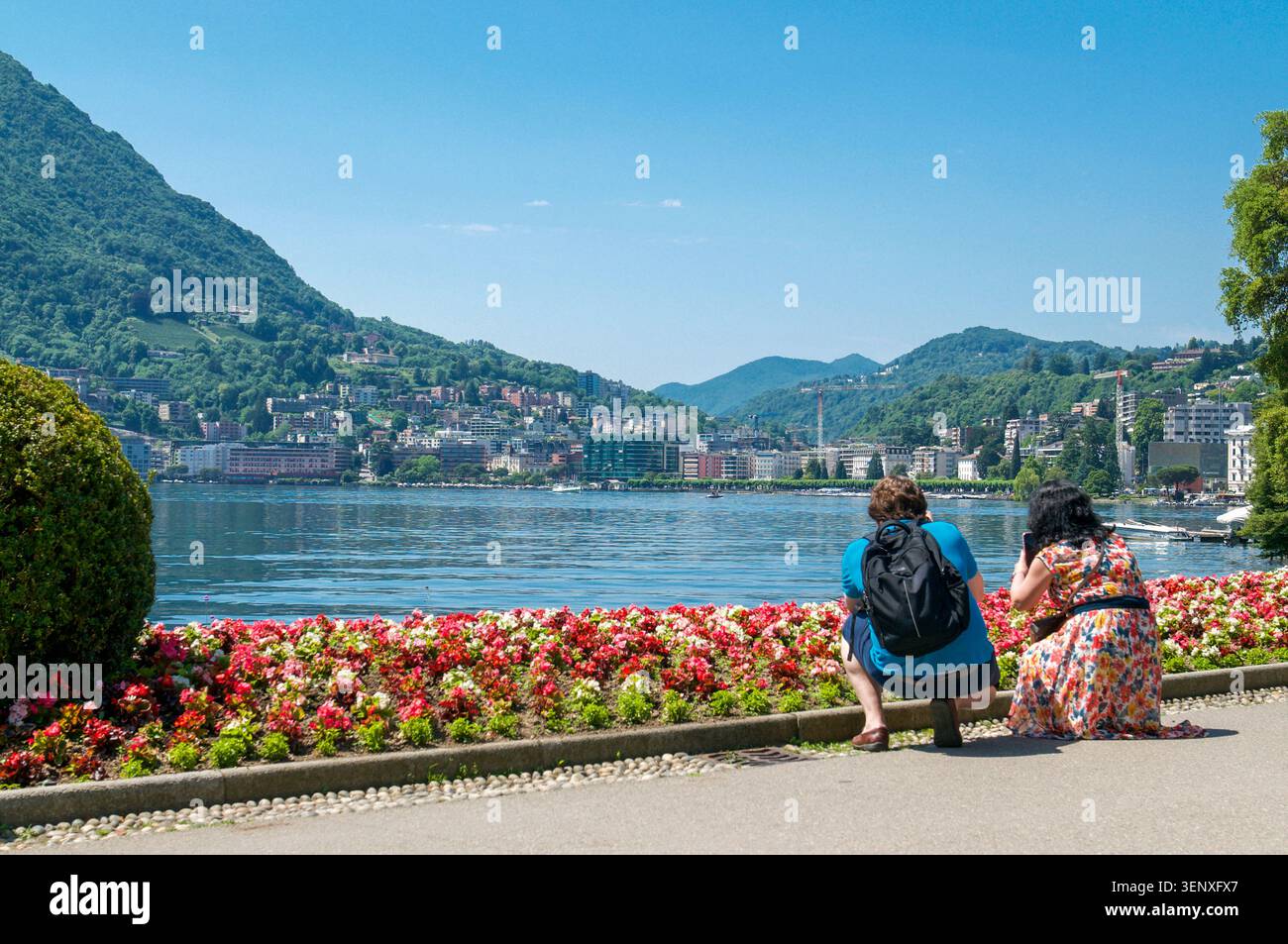 Zwei Touristen von hinten genießen die malerische Aussicht und machen Fotos vom Luganer See und den Bergen vom Parco Ciani Public Park in einem sonnigen Sommer Stockfoto