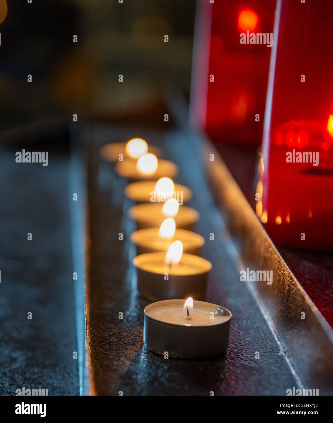 Beleuchtete Kerzen, in parallelen Reihen entlang einer dunklen Metall- oder Holzoberfläche angeordnet, typisch für Kirchenkerzenhalter. Stockfoto