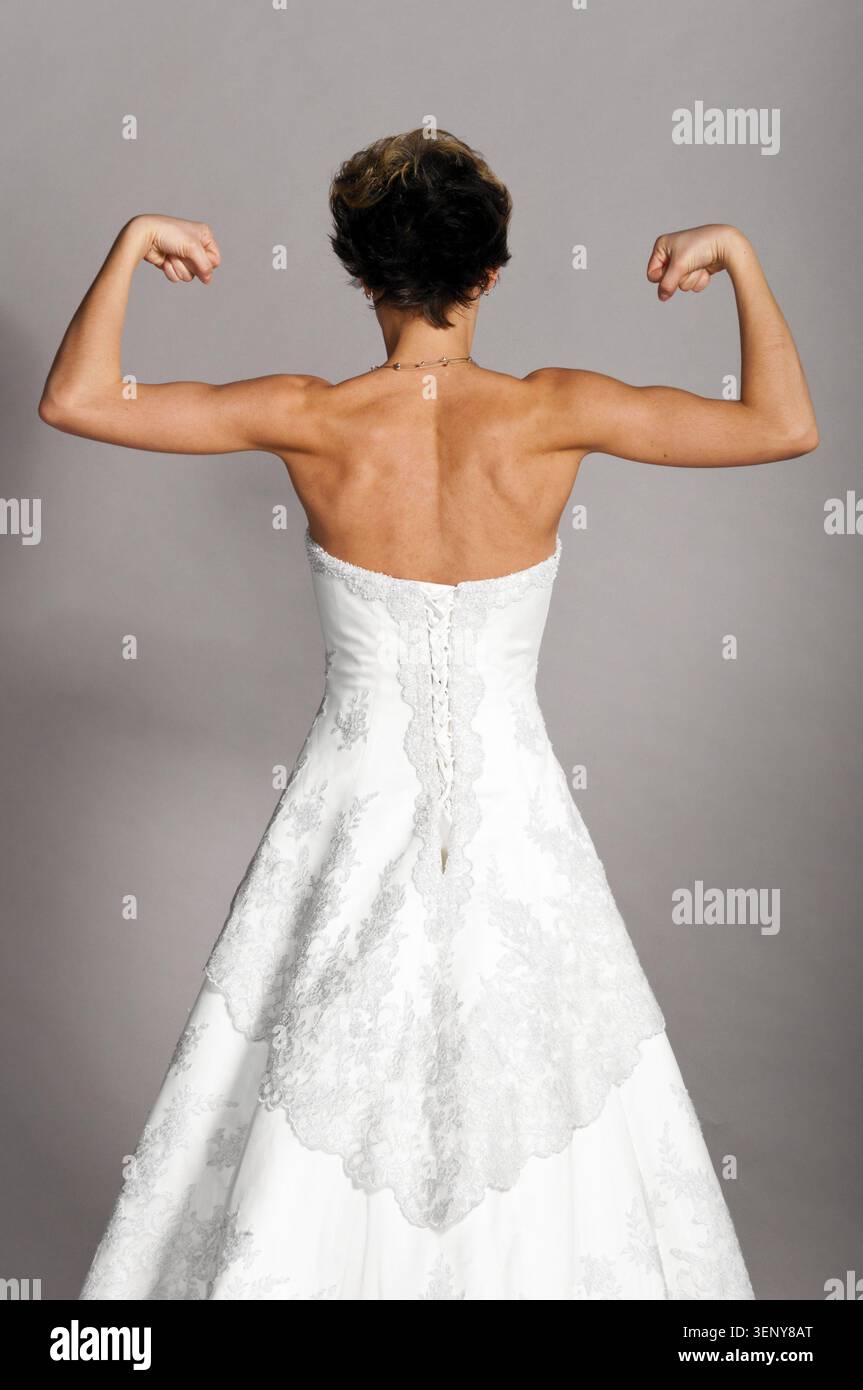 Die schöne Braut steht und zeigt ihre starken Hände im Hochzeitskleid auf grauem Hintergrund, Blick von hinten Stockfoto