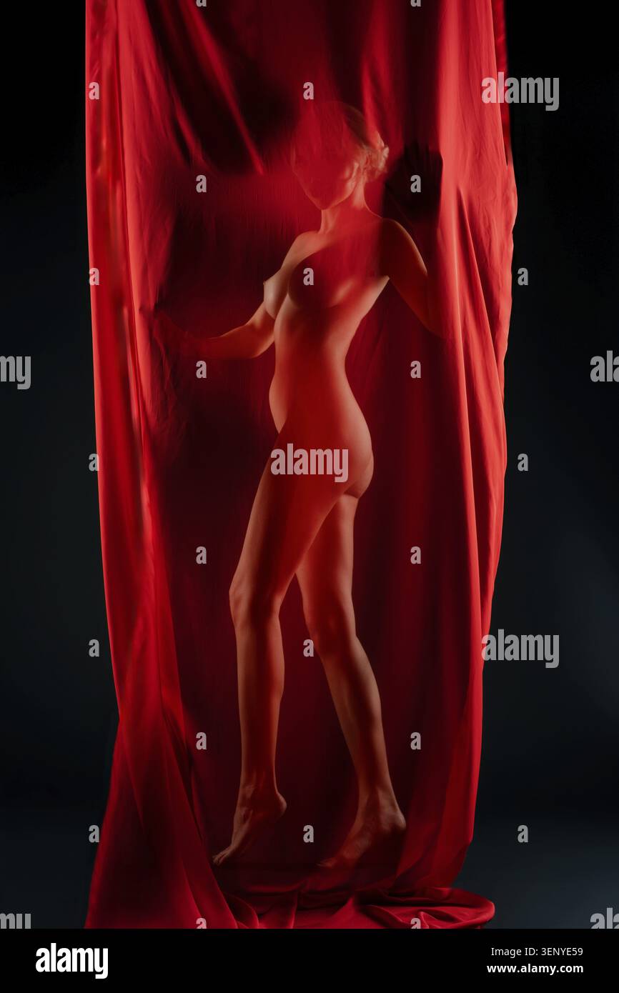 Ein weibliches Model posiert anmutig hinter einem auffälligen roten Tuch in einer Studioumgebung. Die kontrastierenden Farben betonen ihre Silhouette und schaffen eine eth Stockfoto