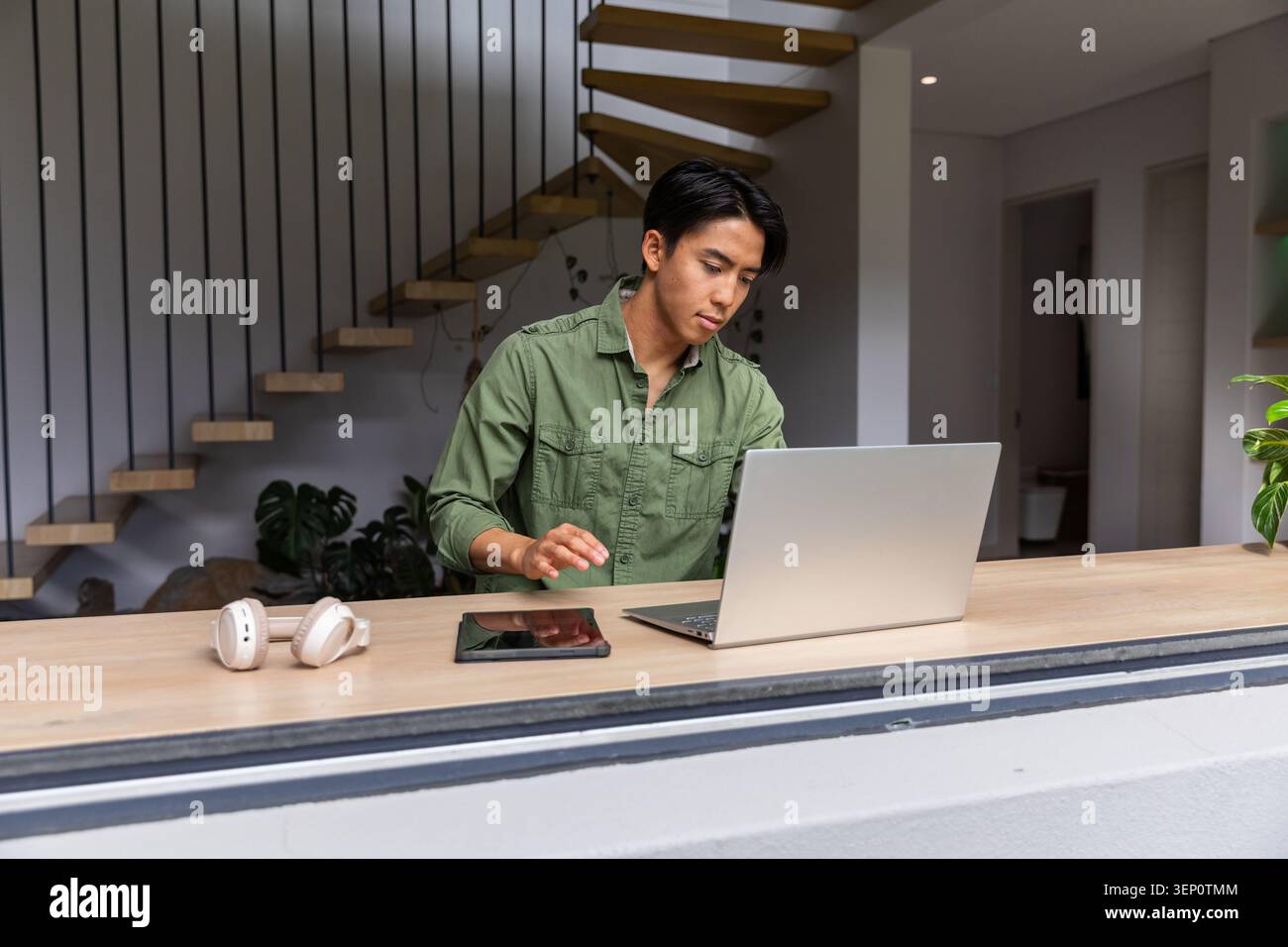 Asiatischer Mann, der hinter Holztheke zu Hause steht, mit silbernem Laptop, Tablet, Headset, in grünem Hemd Stockfoto