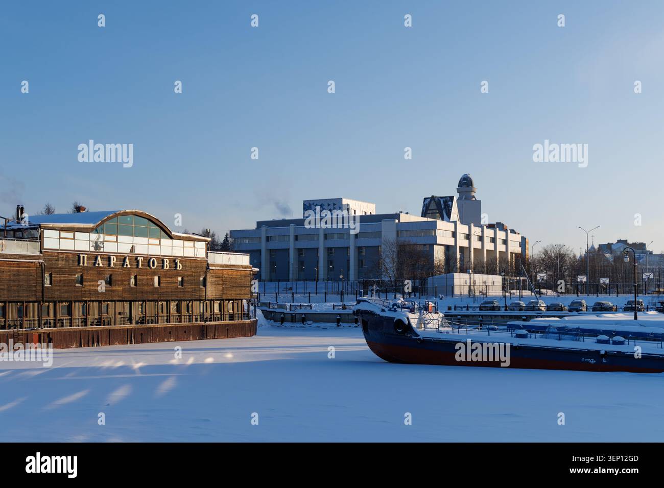 Dieses Café mit Panoramafenstern überblickt die schneebedeckten Ufer der Newa. Ein Boot liegt in der Nähe, und moderne Gebäude stehen in der Ferne Stockfoto