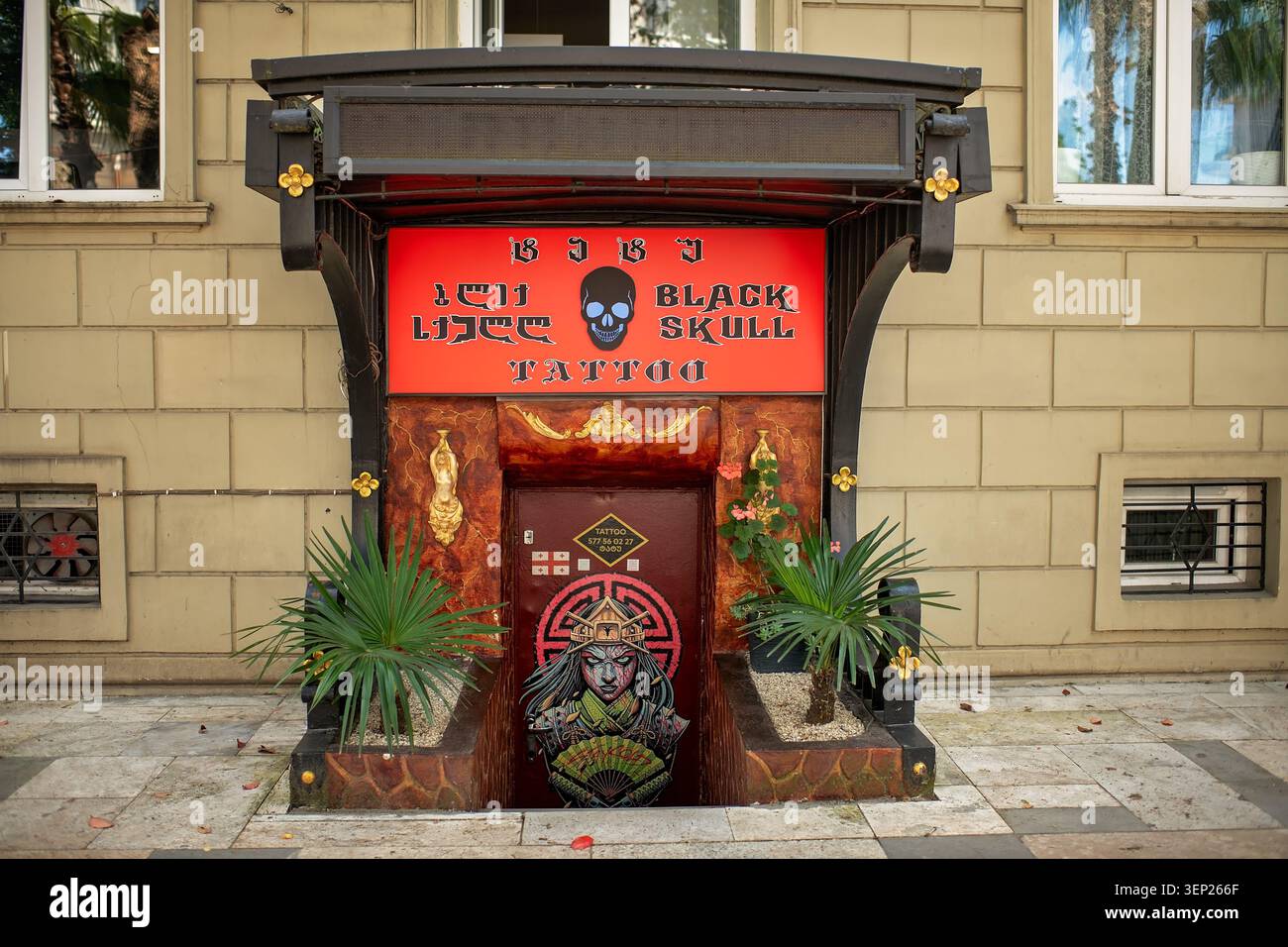 Batumi, Georgia – 9. Juni 2026: Ein stimmungsvoller Eingang zum Tätowiersalon mit einem roten Schild mit Schädelbild, Design an der Tür und kleinen Palmen Stockfoto