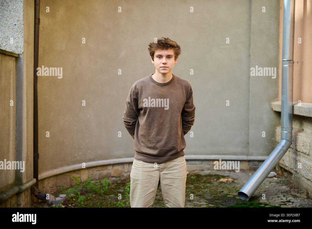 Das junge Individuum lehnt sich kontemplativ an eine strukturierte Wand. Teen steht ruhig mit Händen hinter den erdigen Tönen. Jugendliche Person reflektiert friedlich im schattigen Innenhof Stockfoto