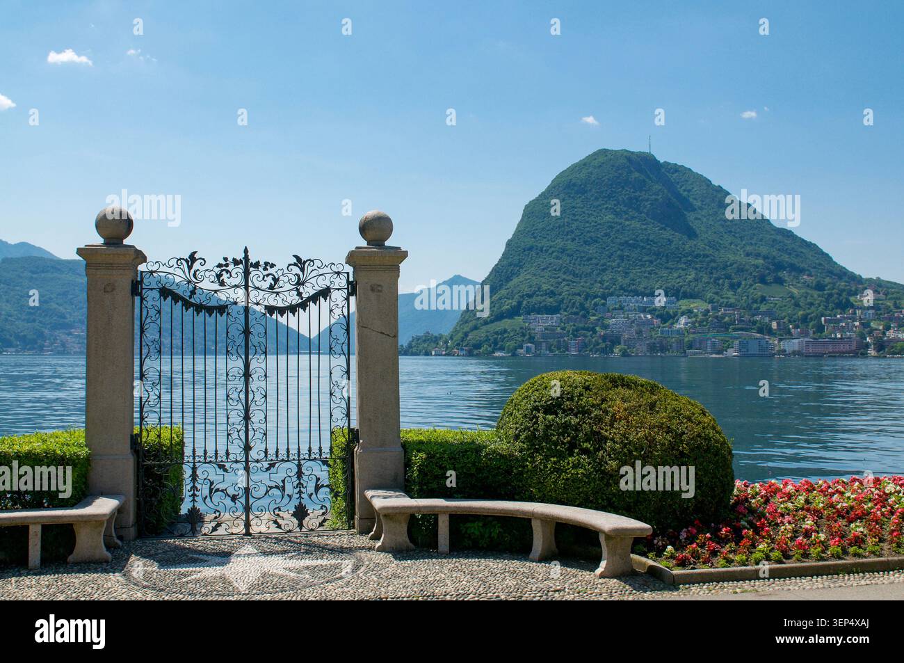 Vertikaler Blick auf das kunstvolle historische Eisentor des Parco Ciani, das sich in Richtung des ruhigen Wassers des Luganer Sees öffnet, mit dem Bergturm des Monte San Salvatore Stockfoto