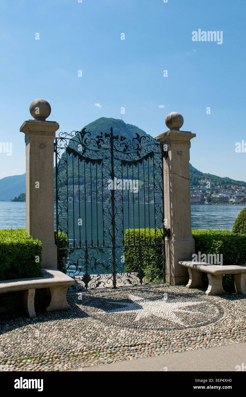 Wunderschöner, vertikaler Blick auf den Luganersee und den Monte San Salvatore Berg durch das kunstvoll verzierte historische schmiedeeiserne Tor des Parco Ciani Public Par Stockfoto