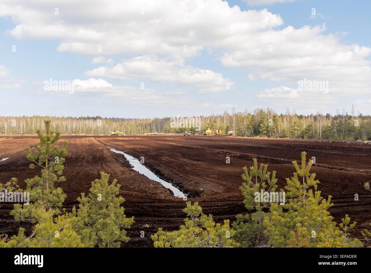 Ein langer Entwässerungsgraben, der durch ein dunkelbraunes Torferntefeld mit Industriebaggern in der Ferne schneidet. Stockfoto