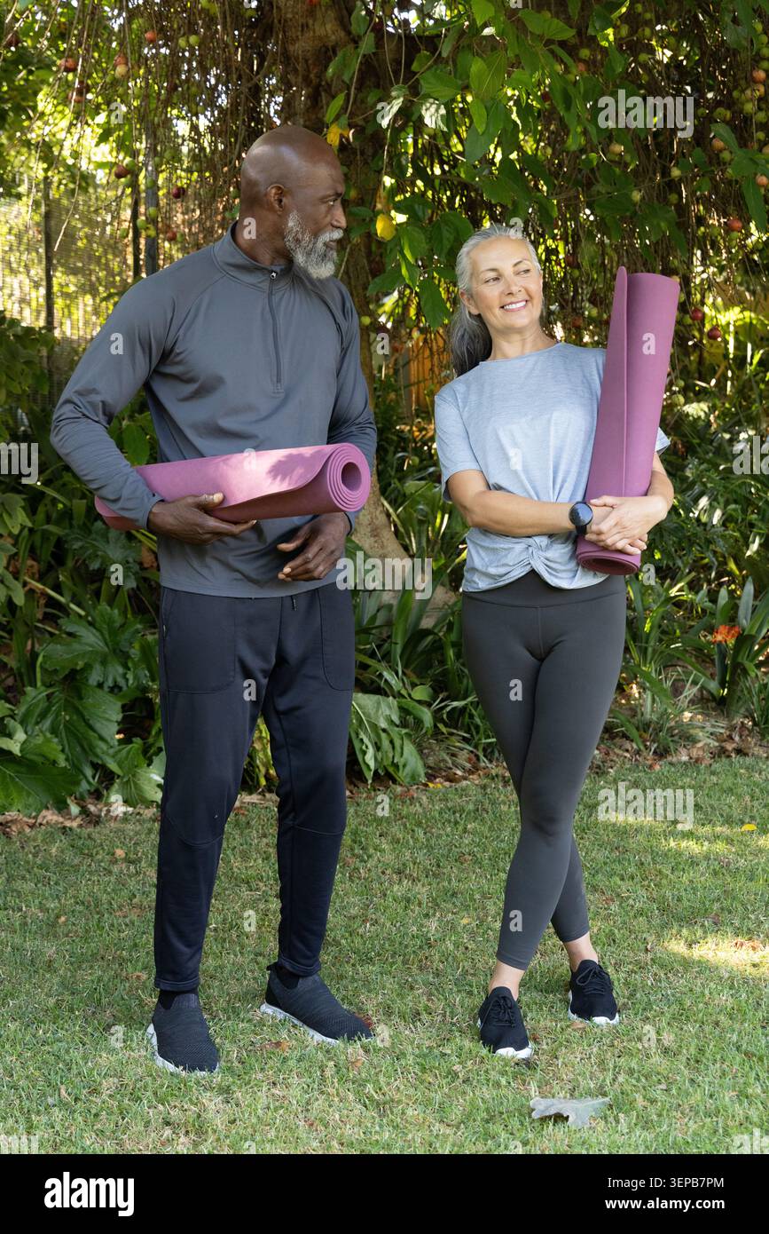 Verschiedene Paare stehen auf dem grasbewachsenen Hof mit gerollten lila Yogamatten, während sie eine Fitnessuhr tragen Stockfoto
