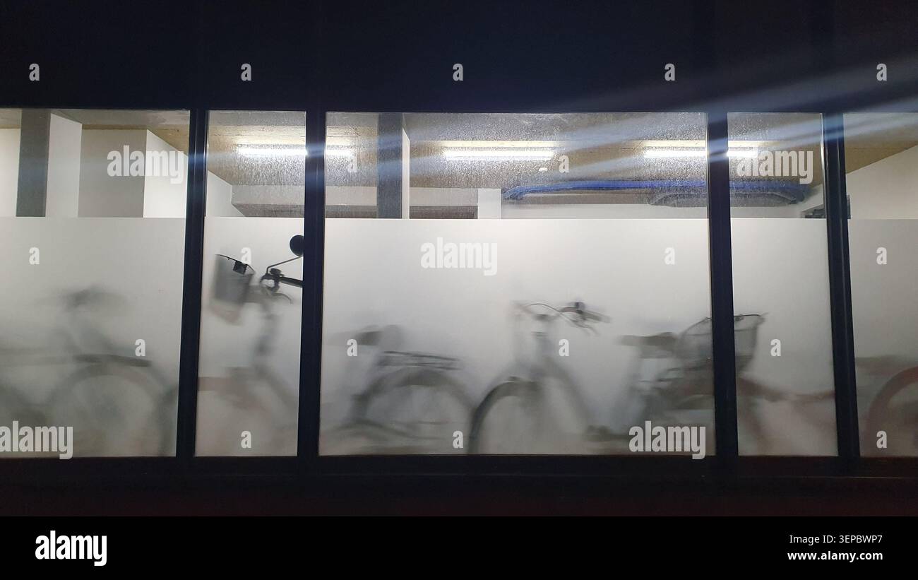 Fahrräder zeichnen weiche Silhouetten hinter mattierten Glaspaneelen in einem Fahrradparkplatz. Bewegungsunschärfe und stimmungsvolle Beleuchtung sorgen für eine ruhige, urbane Atmosphäre Stockfoto