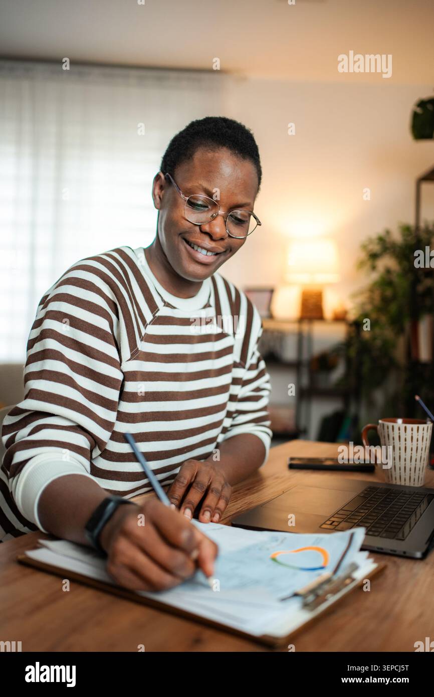 Lächelnde junge Erwachsene Frau, die Hausaufgaben am Schreibtisch schreibt Stockfoto