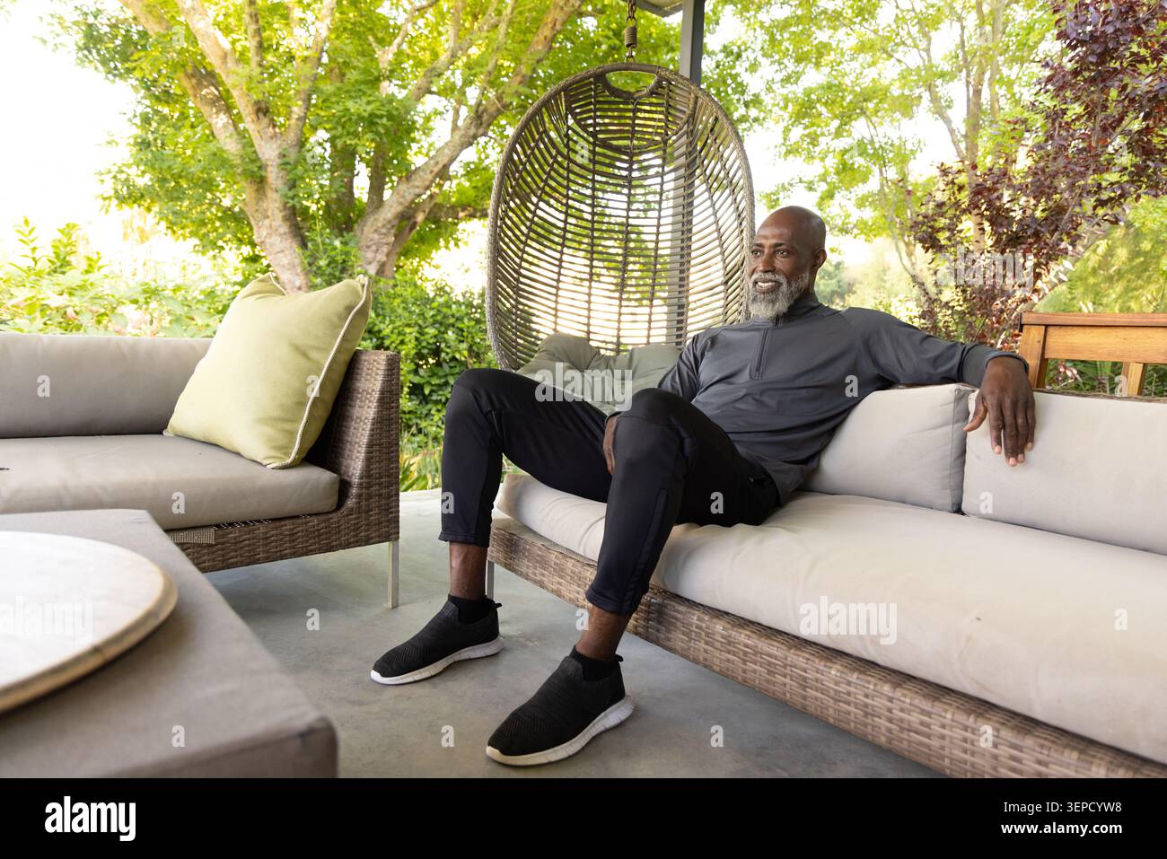 Ranghoher afroamerikanischer Mann, der auf Rattan-Veranda-Sofa in dunklem Pullover neben hängendem Eierstuhl sitzt Stockfoto