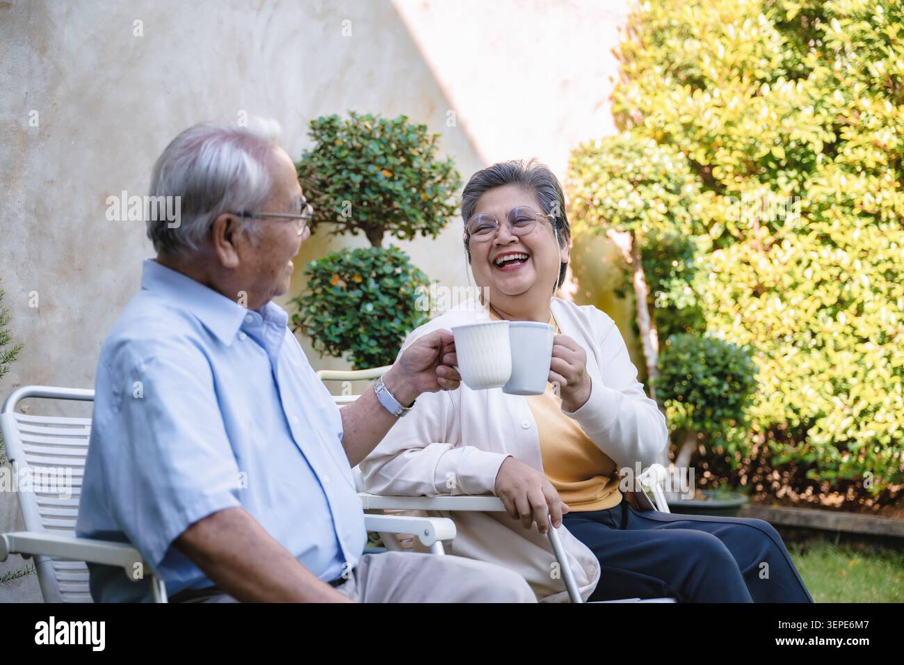 Ältere Ehepaare sitzen in Gartenstühlen und genießen einander Gesellschaft. Der Mann hält eine weiße Teetasse, die Frau lächelte ihn warm an. Sie teilen einen friedlichen Moment mit mir Stockfoto