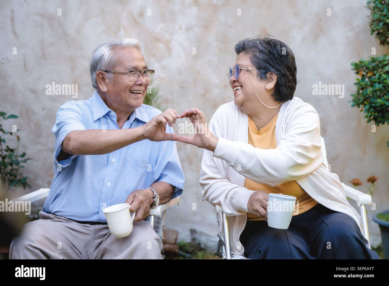 Älteres Ehepaar sitzt in Gartenstühlen. Jeder hält eine weiße Teetasse. Sie lächeln und verbinden sich herzförmig mit den Händen. Ihre Freude ist klar und ehrlich. Ideal für Stockfoto