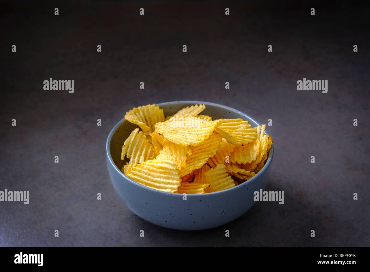 Goldene, wellige Chips füllen eine gesprenkelte Schüssel. Dunkle Oberfläche steht im Kontrast zu knusprigem, einladendem Snack. Die einfache Zusammensetzung unterstreicht die Textur und Farbe des Snacks. I Stockfoto