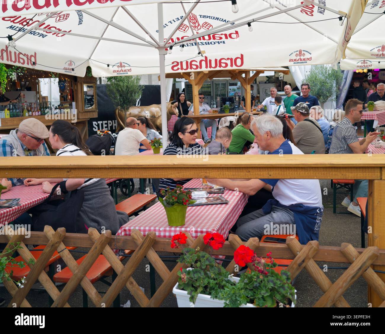 Pforzheim Deutschland - 14. Juni 2026 Machen Sie eine geschäftige Freiluft-Biergarten-Szene mit Gruppen von Personen, die an hölzernen Picknicktischen sitzen, die mit Rot-weiß bedeckt sind Stockfoto