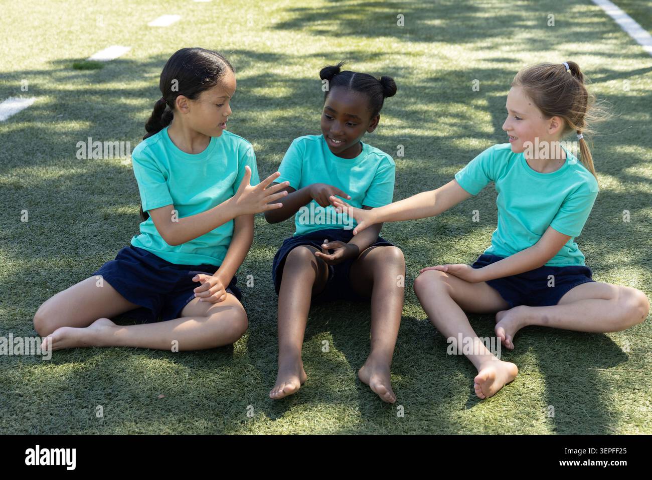 Verschiedene Schulmädchen sitzen barfuß auf dem Rasen in passenden blauen Hemden marineblauen Shorts und spielen Handspiele Stockfoto