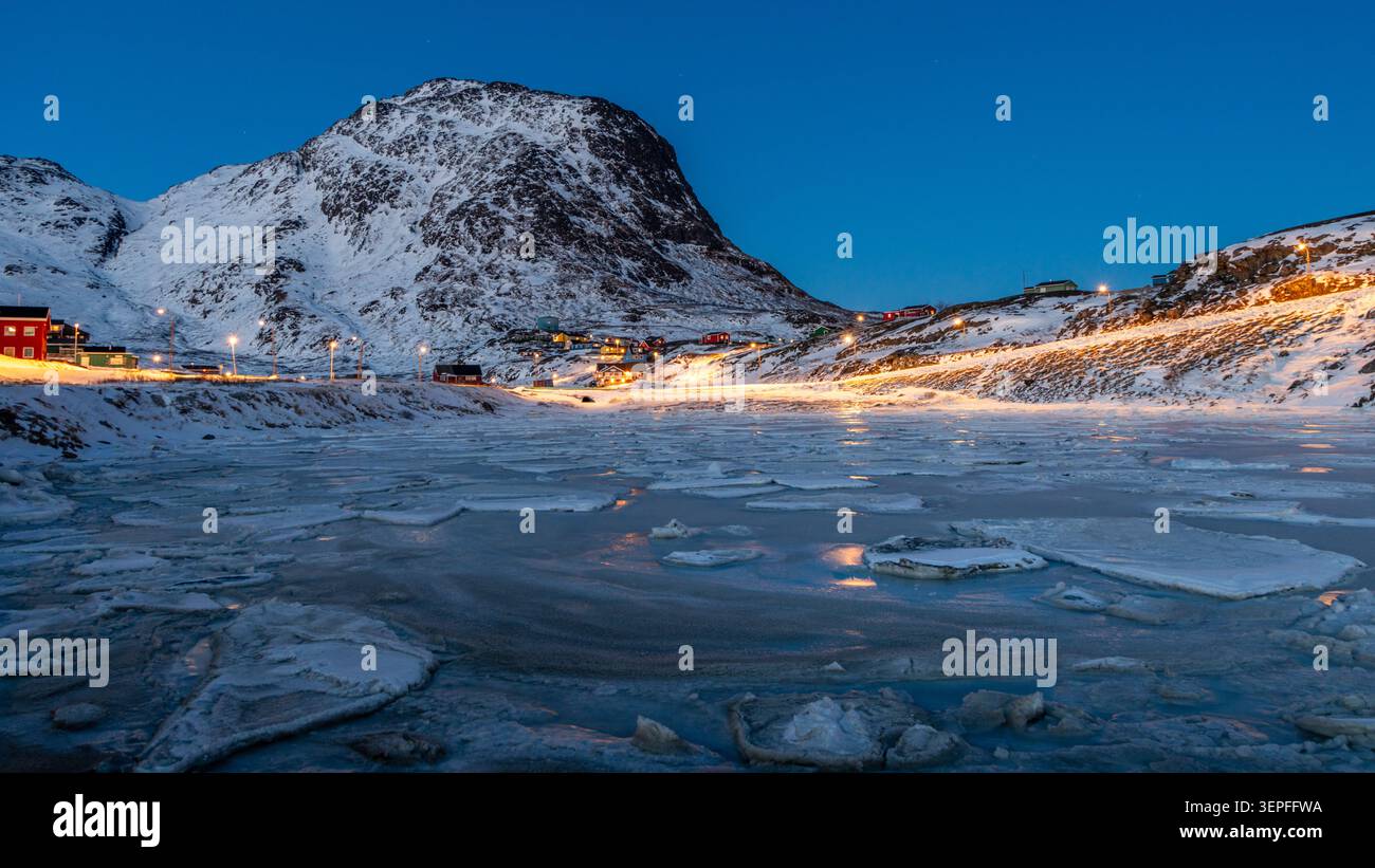 Winterabendlandschaft von Narsaq mit gefrorenem Wasser, schneebedeckten Bergen, Straßenlaternen und arktischer Dämmerung, Grönland Stockfoto