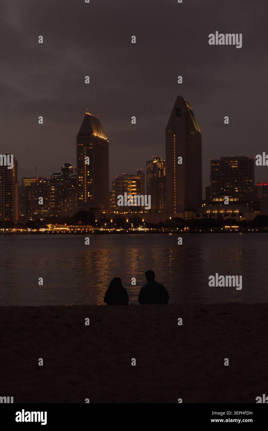 Nächtliche Skyline über ruhiges Wasser mit zwei Personen, die am Strand sitzen und in der Abenddämmerung ein ruhiges urbanes Uferviertel zeigen Stockfoto