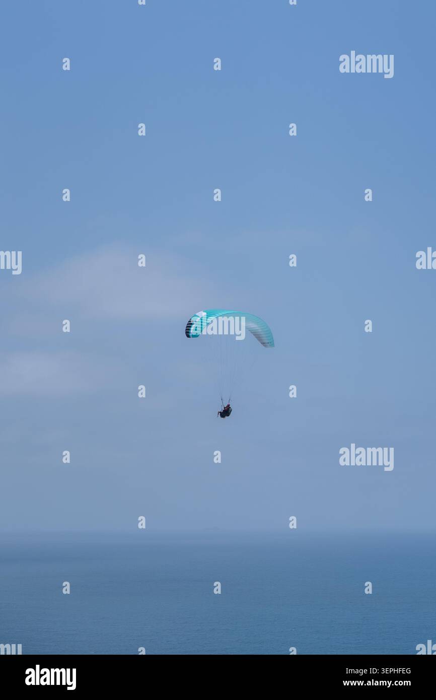 Ein einsamer Gleitschirmflieger, der hoch über dem ruhigen Meer unter einem klaren blauen Himmel gleitet, mit viel freiem Raum und einem friedlichen, luftigen Gefühl. Stockfoto