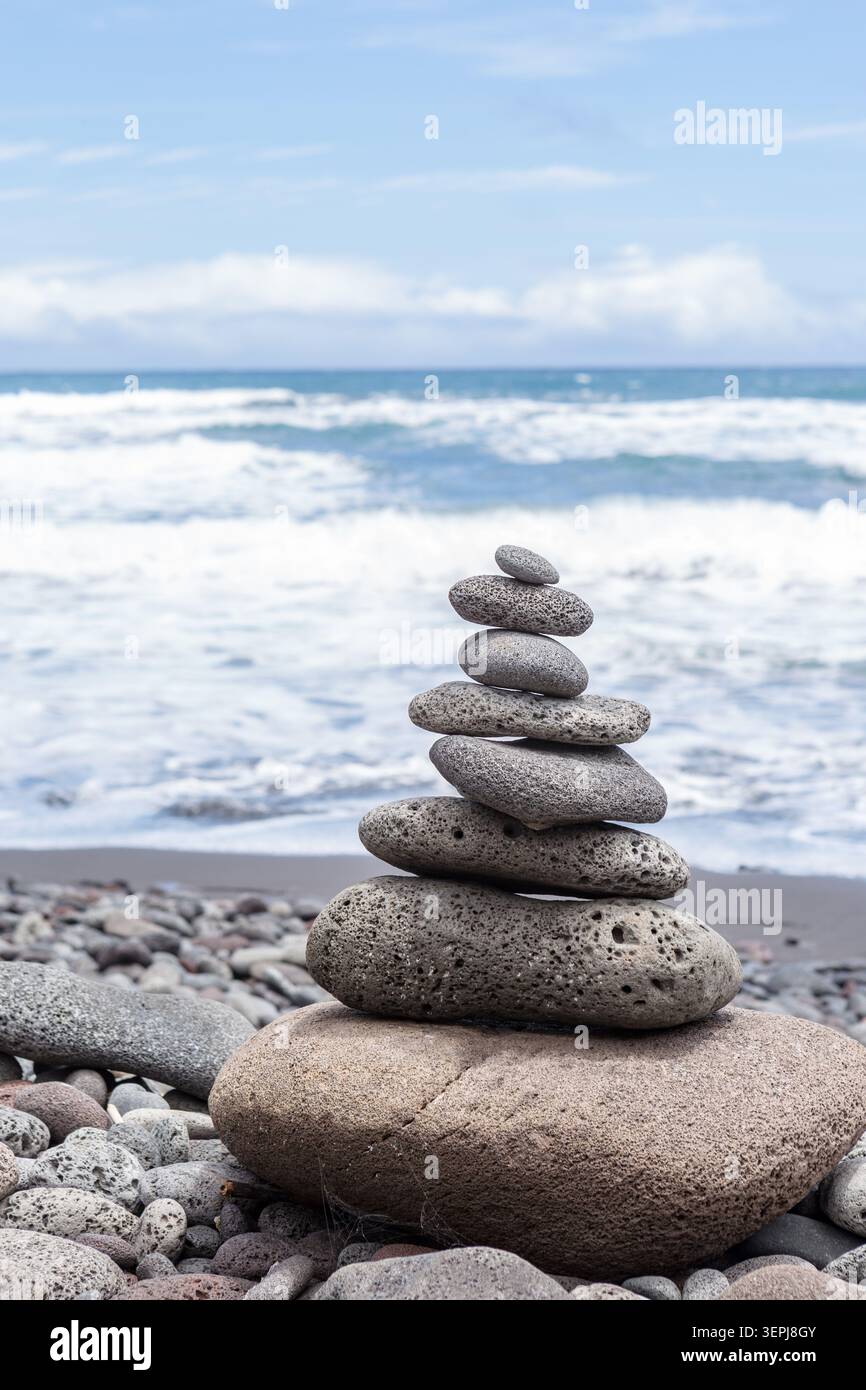 Ausgeglichener vulkanischer Steinkairn an einem steinigen Strand mit Wellen des Pazifischen Ozeans im Hintergrund auf Hawaiis Big Island, symbolisiert Gleichgewicht, Achtsamkeit und . Stockfoto