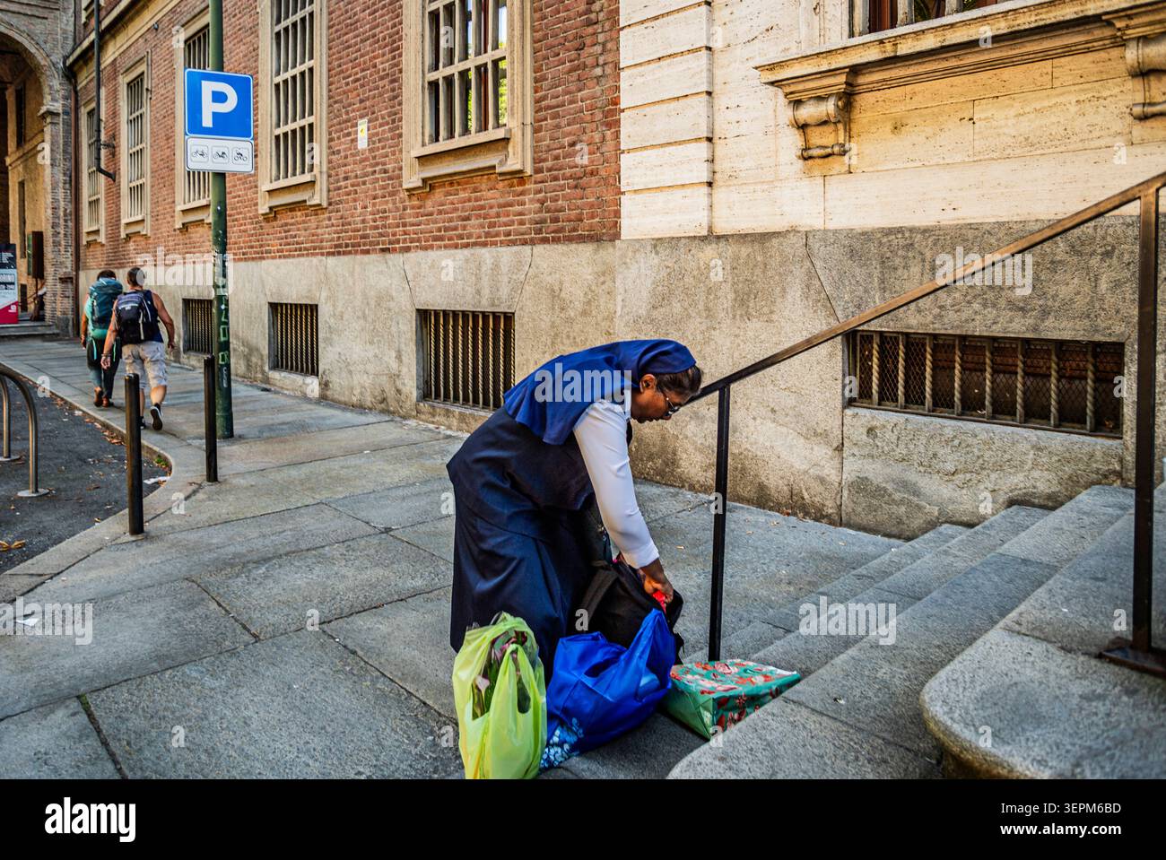 Eine Nonne arrangiert Taschen auf den Stufen eines historischen Gebäudes in Corso Valdocco, Turin, Piemont, Italien, während Fußgänger die Straße im Hintergrund entlang laufen. Stockfoto