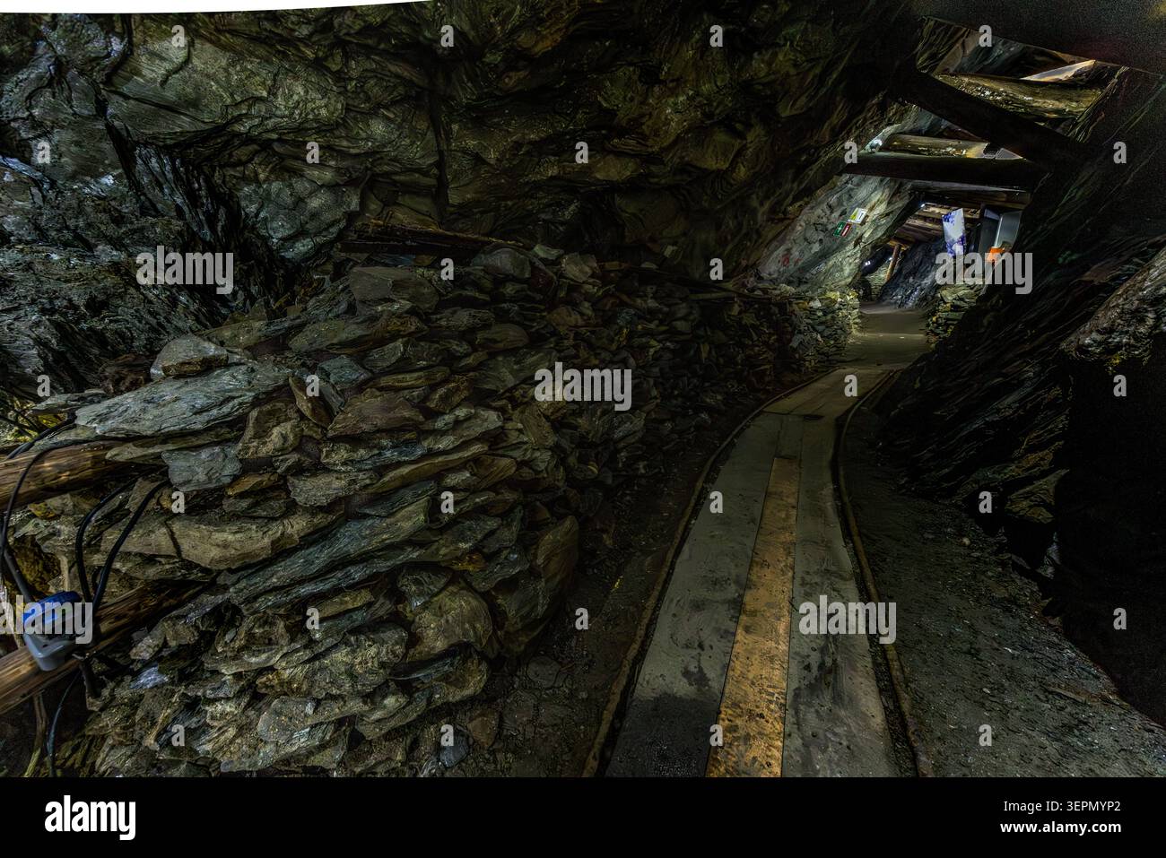 Eine von Hand gelegte Trockenmauer stützt die Tunnelmauer in der Hochfeld-Ausstellungsmine, während ein Holzsteg tiefer in die ehemalige Kupfermine im Untersulzbachtal führt. Am Ende des beleuchteten Gangs ist ein Abzweigstunnel zu sehen. Rosental, Neukirchen am Großvenediger, Bezirk Zell am See, Salzburg, 5741, Österreich Stockfoto