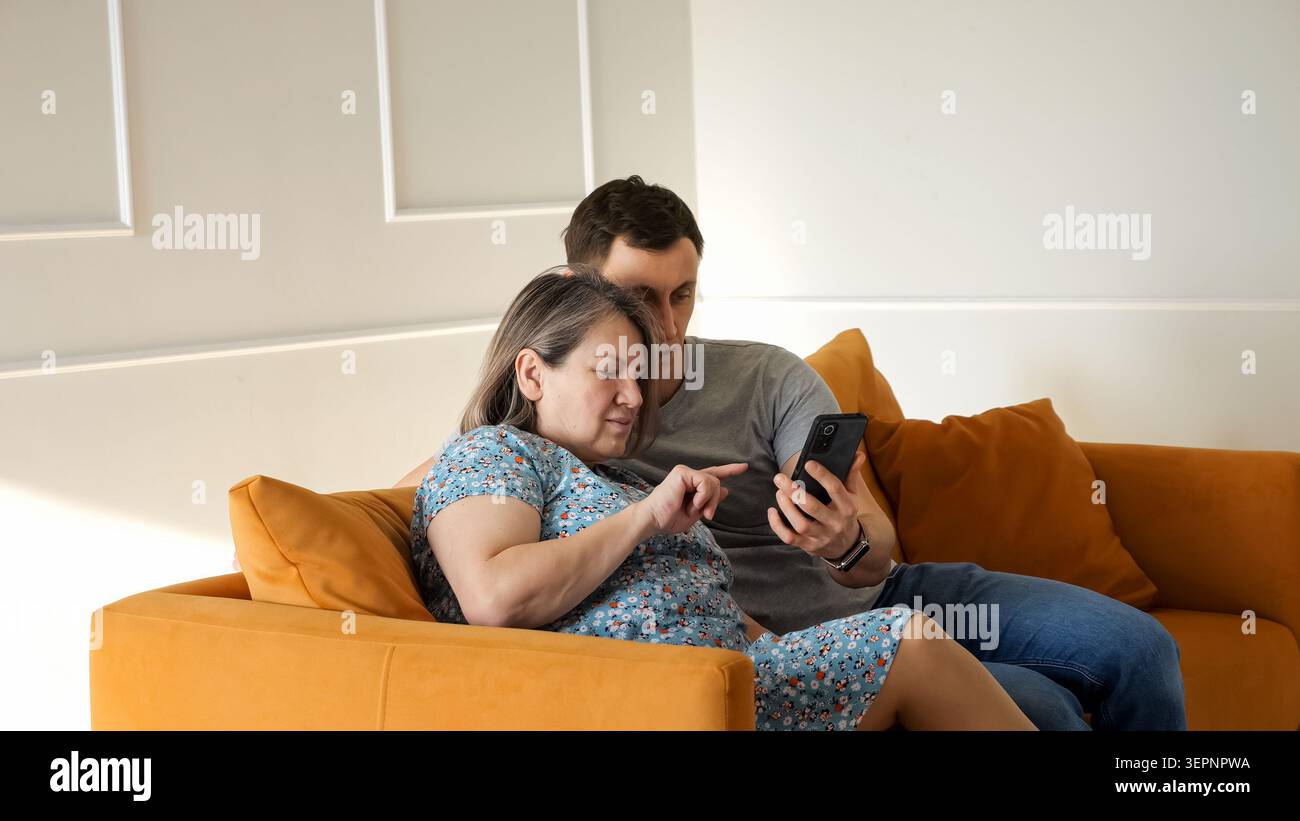 Der Sohn hilft seiner älteren Mutter, neue Technologien und Computerfähigkeiten auf einem Laptop zu erlernen, während er sich auf einem gemütlichen Sofa in einem modernen Wohnzimmer entspannt Stockfoto