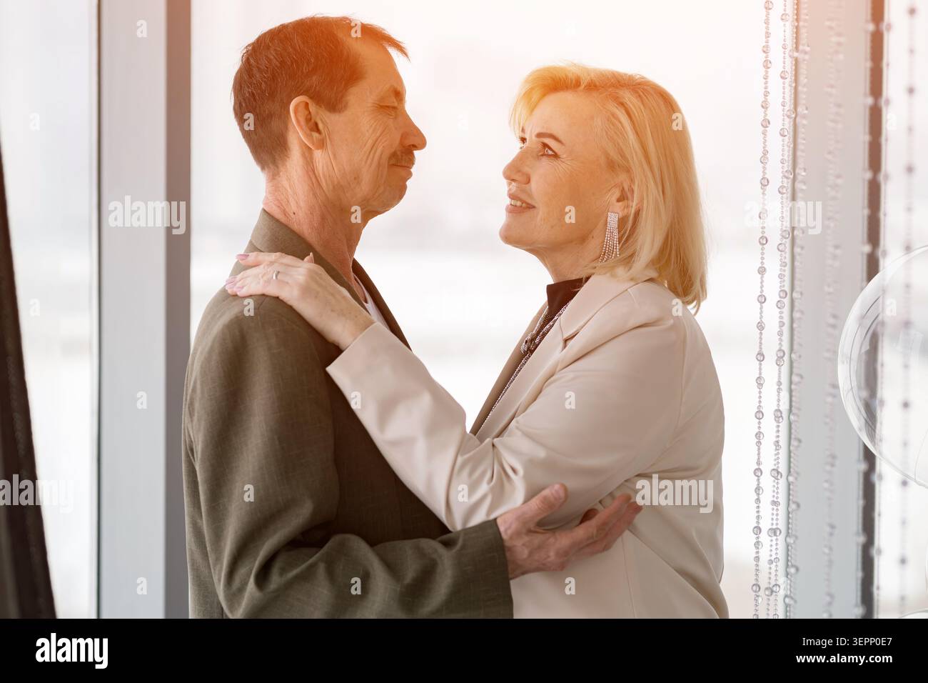 Weißes Paar Umarmen Sich Durch Ein Sonnendurchflutetes Fenster, Reife Partner Feiern Jubiläum Im Modernen Glasbüro, Warme Hintergrundlichter Sanftes Lächeln Und Hände Stockfoto