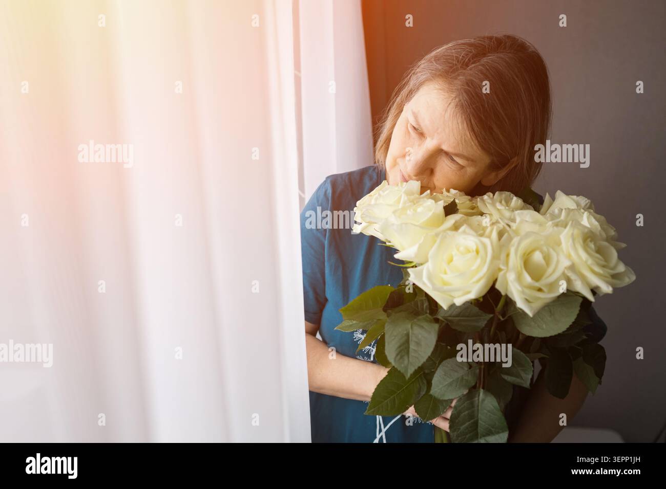 Frau riecht einen Strauß weißer Rosen an einem hellen Fenster in sanftem Morgenlicht Stockfoto