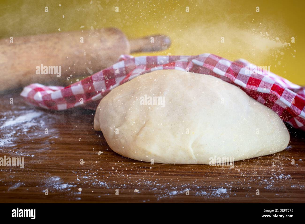 Frischer, roher Hefeteig, der unter einem karierten Küchentuch auf einem Holzbrett mit einer Nudelnadel und Mehlstaub aufsteigt Stockfoto