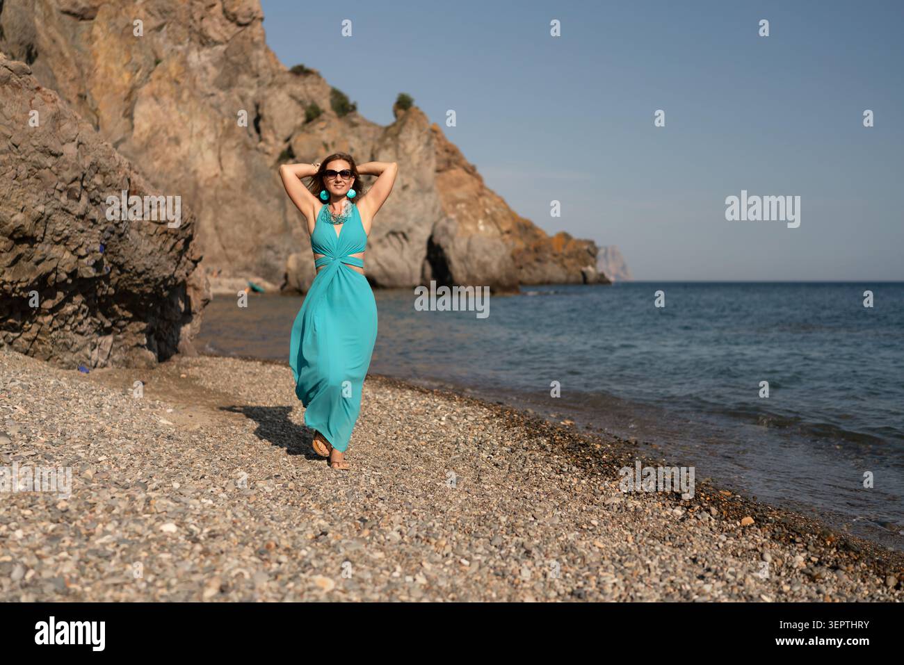 Frau, Strand, Sommer: Lächelnde Frau in türkisfarbenem Kleid spaziert am Kiesstrand an den Klippen des Ozeans unter klarem blauem Himmel. Stockfoto