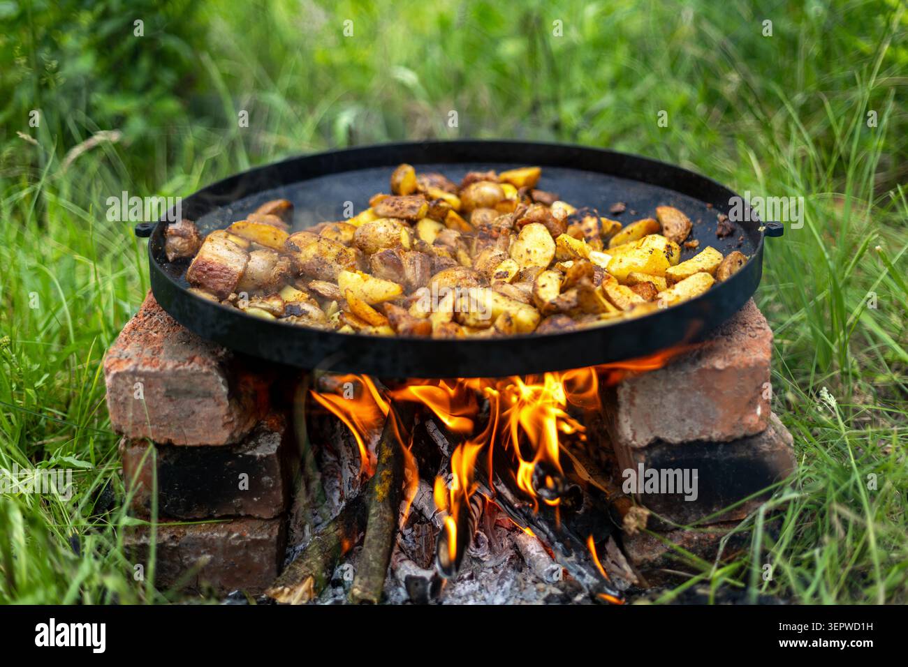Kartoffeln und Fleisch über offenem Feuer braten. In Scheiben geschnittene Kartoffeln und Fleischstücke in einer großen schwarzen Pfanne über hellen Flammen braten, wobei Rauch aus A steigt Stockfoto