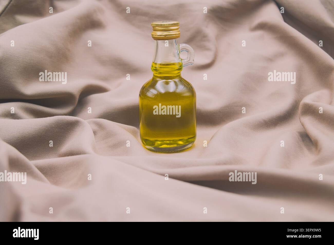 Eine kleine Glasflasche mit goldenem Olivenöl, elegant inszeniert auf weichen, neutralen Stofffalten Stockfoto