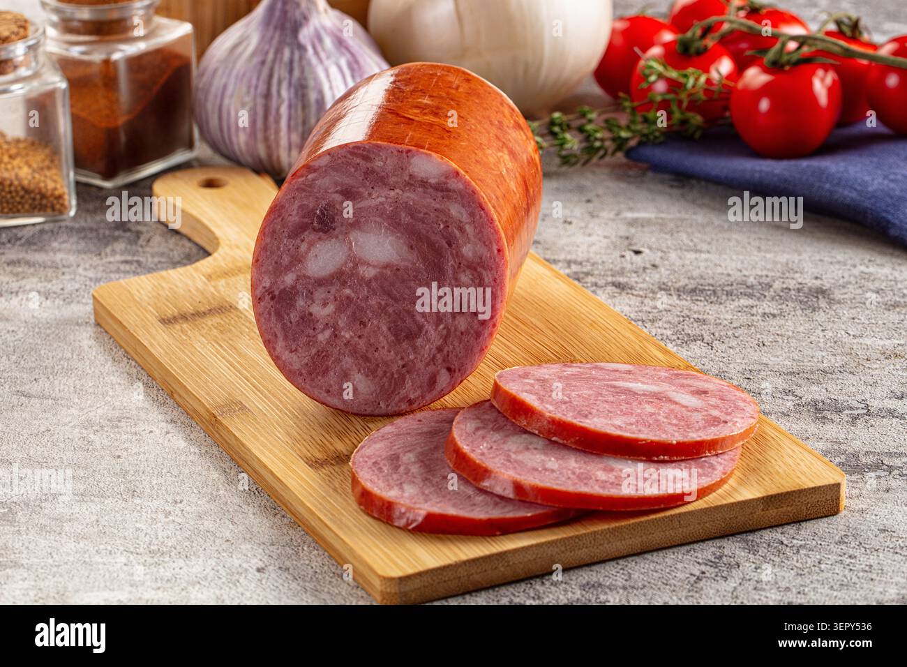 Geräucherter Wurststamm in runde Stücke geschnitten auf hölzernem Schneidebrett, umgeben von frischen Tomaten, Knoblauch und Gewürzgläsern auf Steinoberfläche Stockfoto